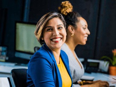 Happy looking office worker sitting at her office desk looks towards the camera, with another staff member sitting at her desk behind her